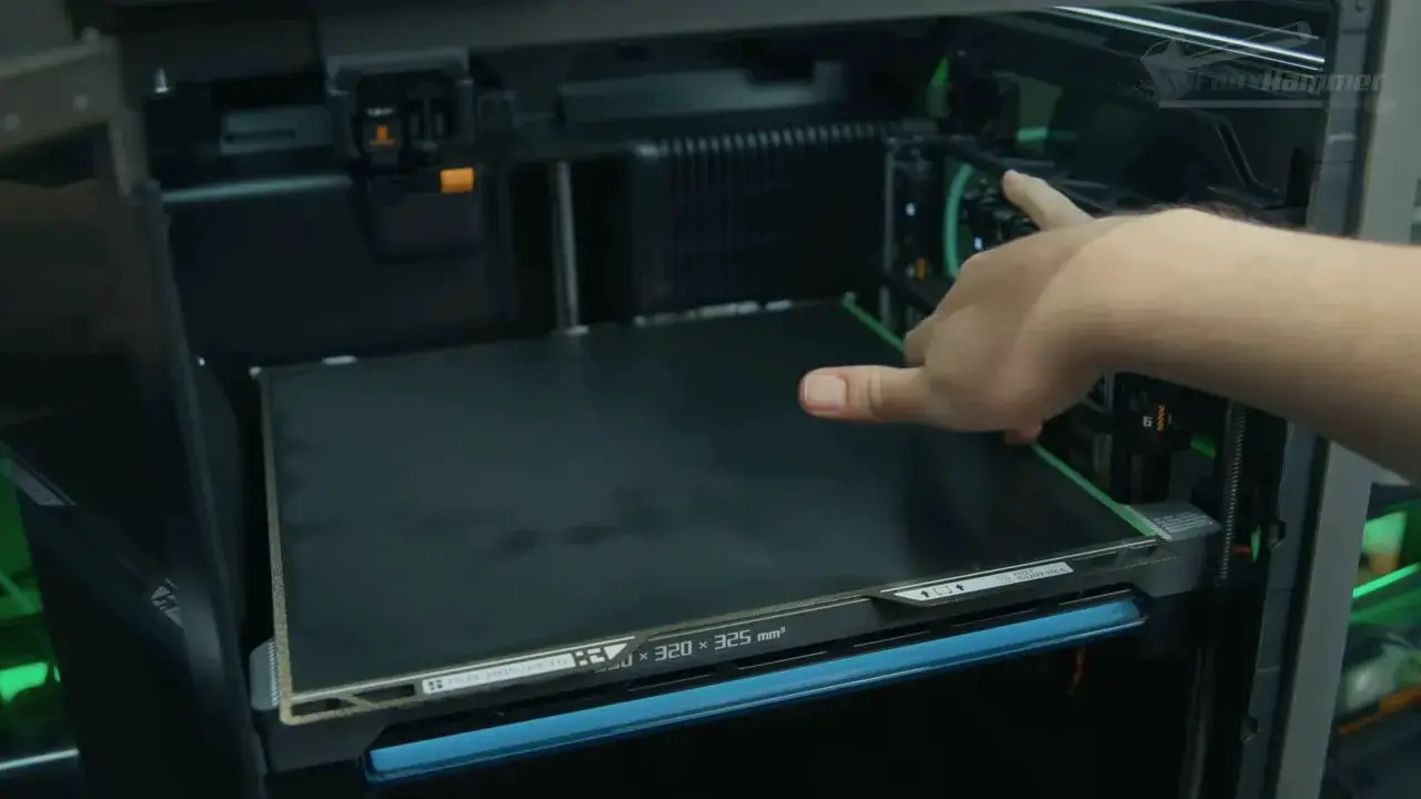 View inside a Bambu H2 enclosed printer chamber with build area overlay and a hand pointing toward the bed
