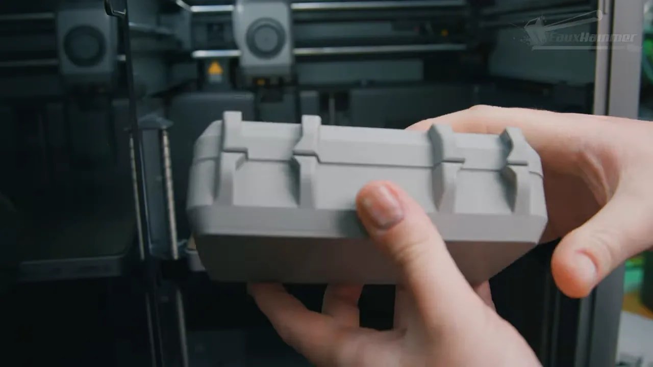 Hands holding a larger white 3D printed part from an enclosed 3D printer