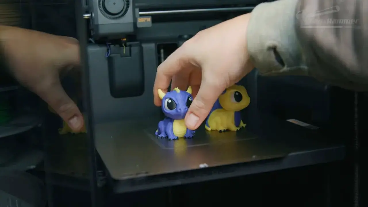 Dual-color figurines on the Bambu X2D build plate with the operator’s hand adjusting the prints