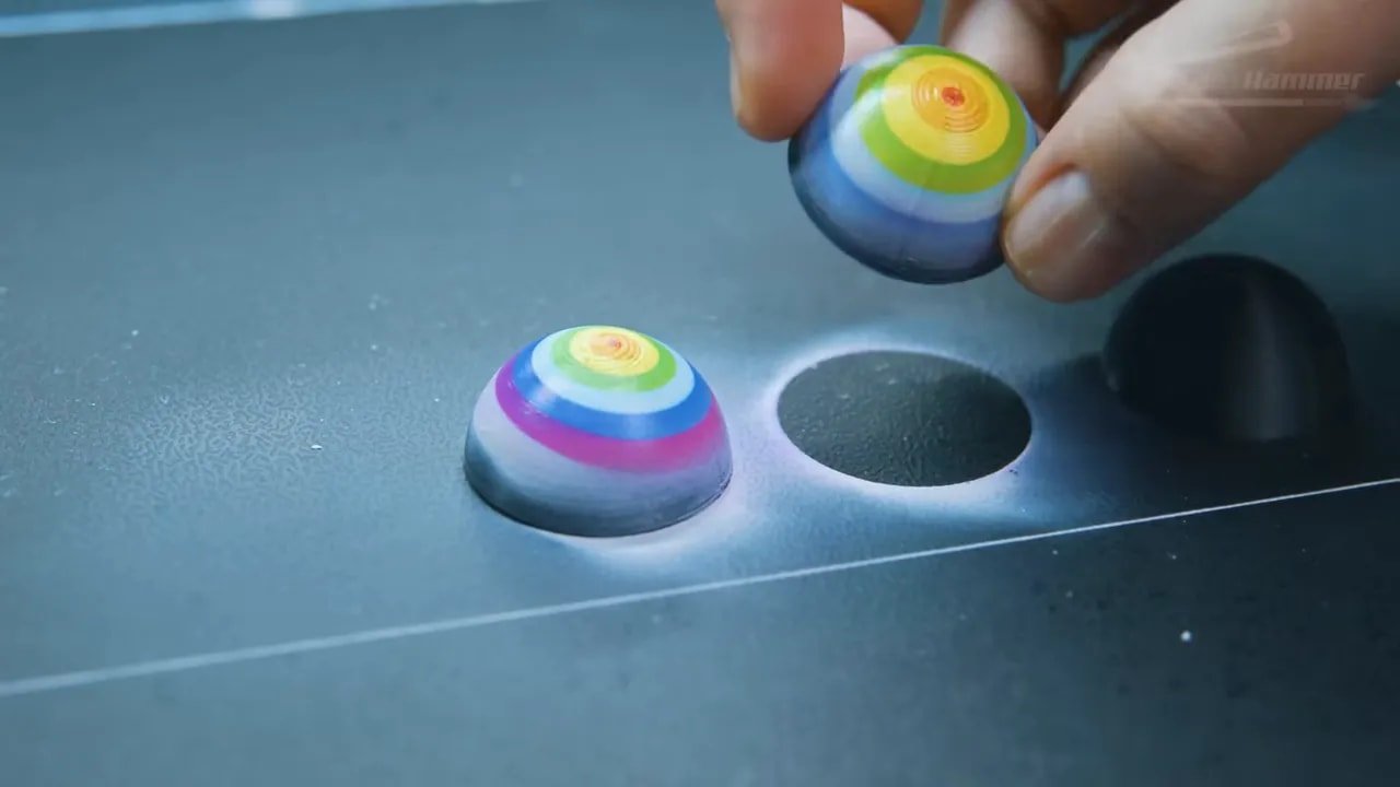 Finished cylindrical cap with rainbow target-style print on a printer bed next to a second test piece