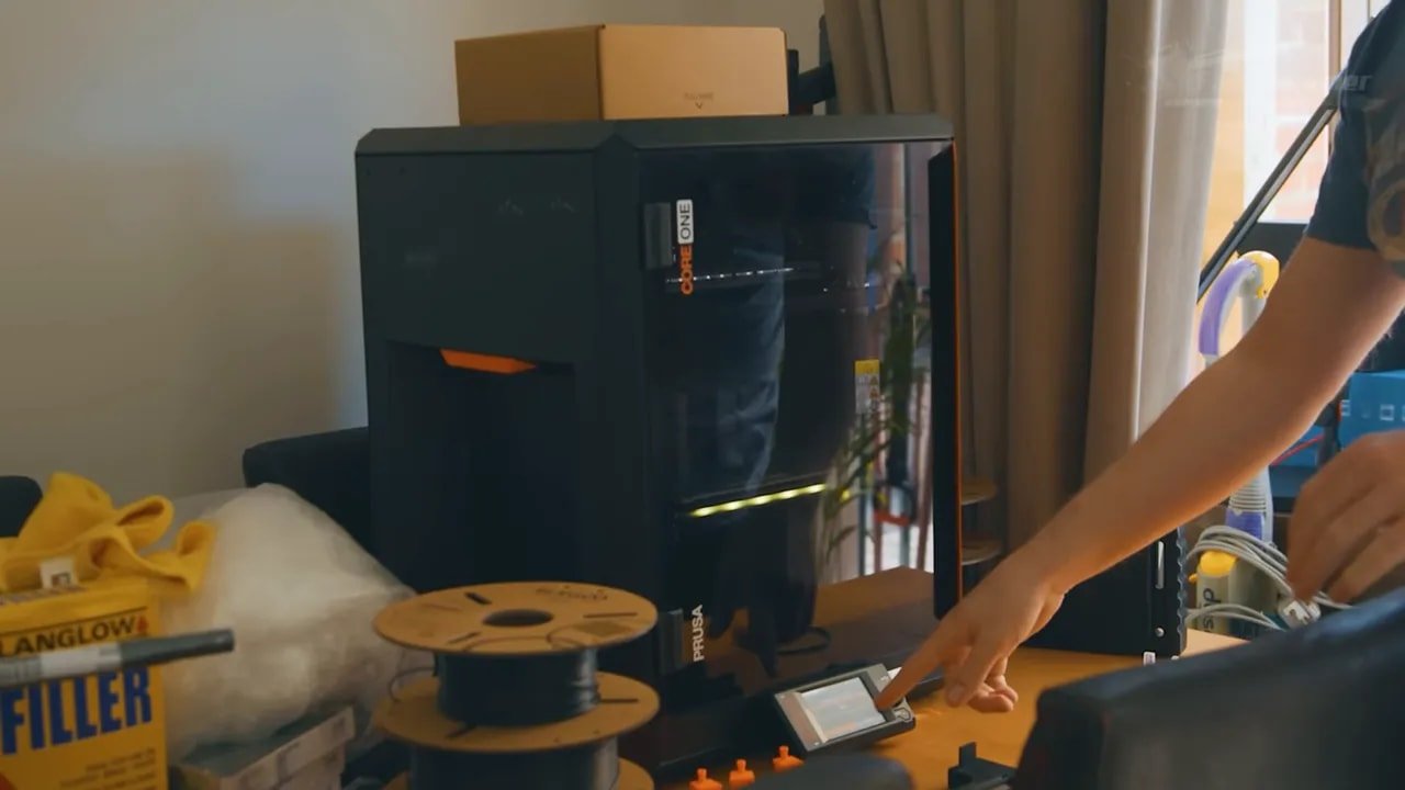 Prusa Core One enclosed printer in a room with the front window visible while a hand adjusts the printer